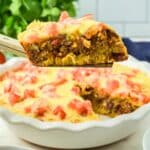 A slice of cheesy, baked taco pie casserole with ground meat and diced tomatoes is lifted from a pie dish containing the remaining casserole.