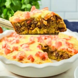 A slice of cheesy, baked taco pie casserole with ground meat and diced tomatoes is lifted from a pie dish containing the remaining casserole.
