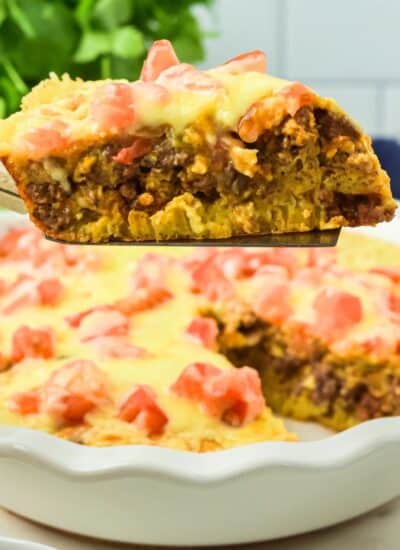 A slice of cheesy, baked taco pie casserole with ground meat and diced tomatoes is lifted from a pie dish containing the remaining casserole.