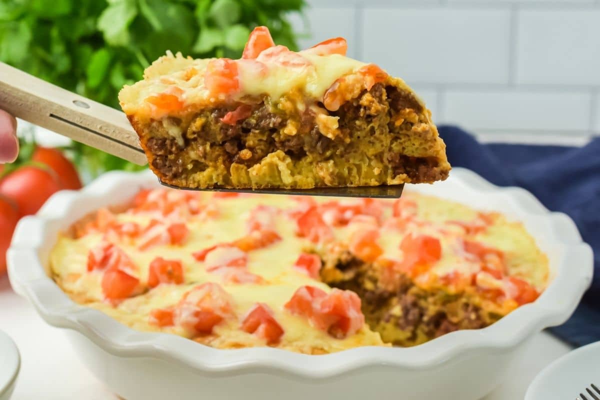 A slice of taco casserole with ground beef, melted cheese, and diced tomatoes—reminiscent of a classic taco pie—is being lifted from a round baking dish.