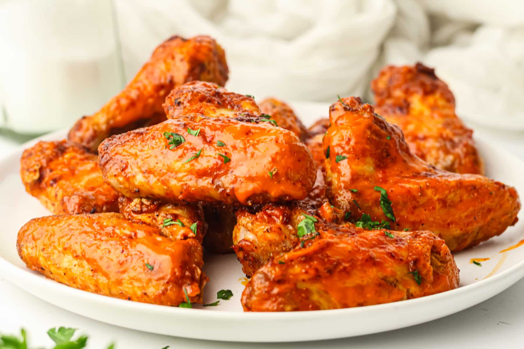 A white plate filled with saucy Air Fryer Frozen Chicken Wings, garnished with chopped herbs, sits on a white surface.
