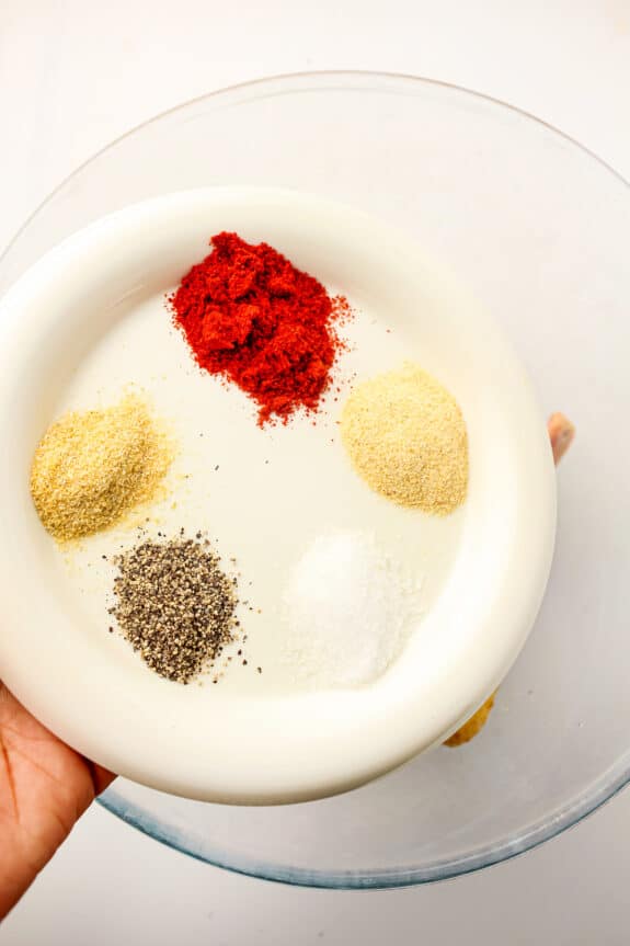A hand holds a white plate with five piles of spices: paprika, garlic powder, onion powder, black pepper, and salt over a glass bowl for Air Fryer Frozen Chicken Wings recipe.