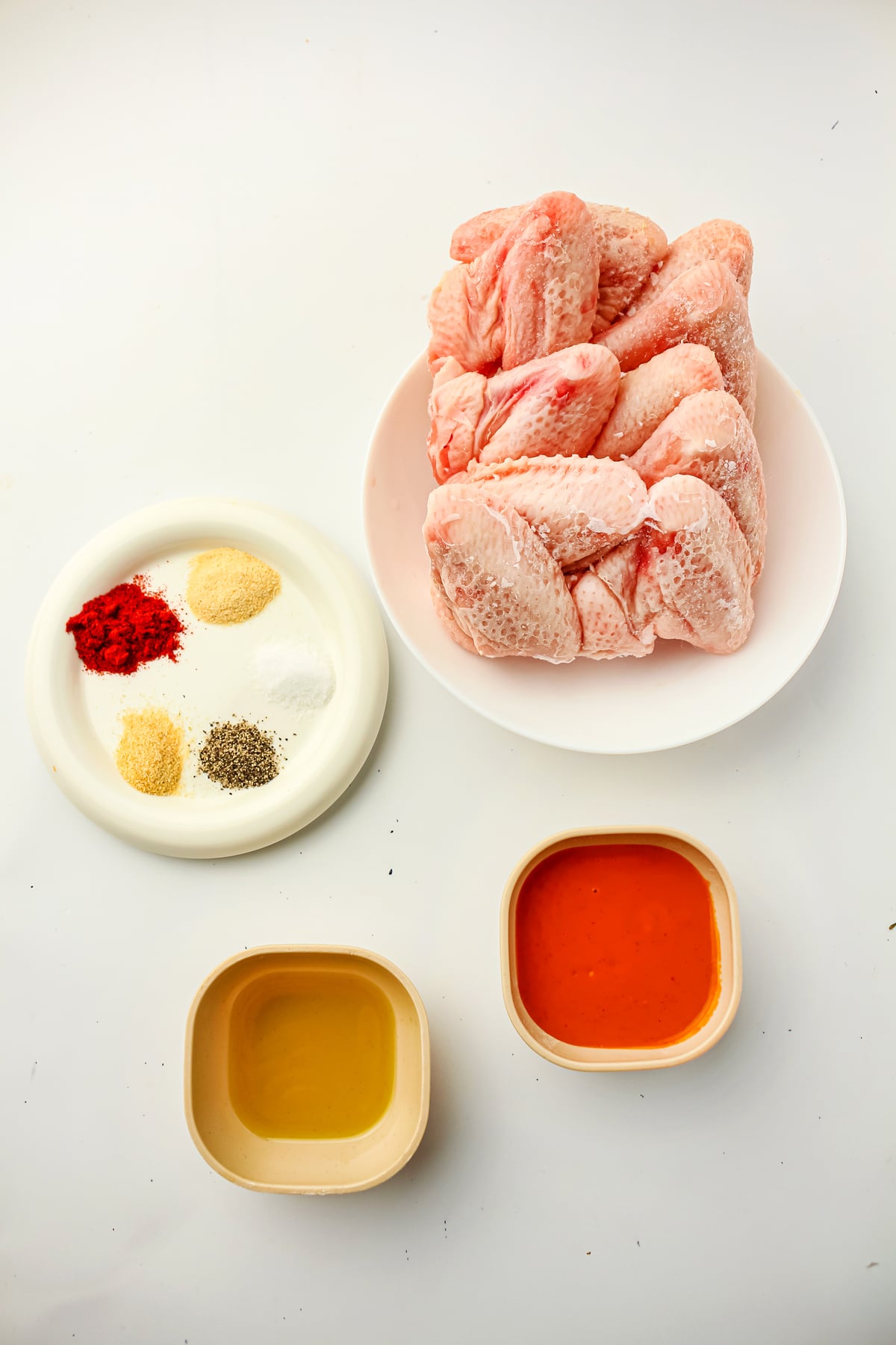Ingredients for Air Fryer Frozen Chicken Wings include a plate of raw frozen chicken wings, avocado or olive oil, garlic powder, smoked paprika, onion powder, black pepper, salt, and buffalo sauce.