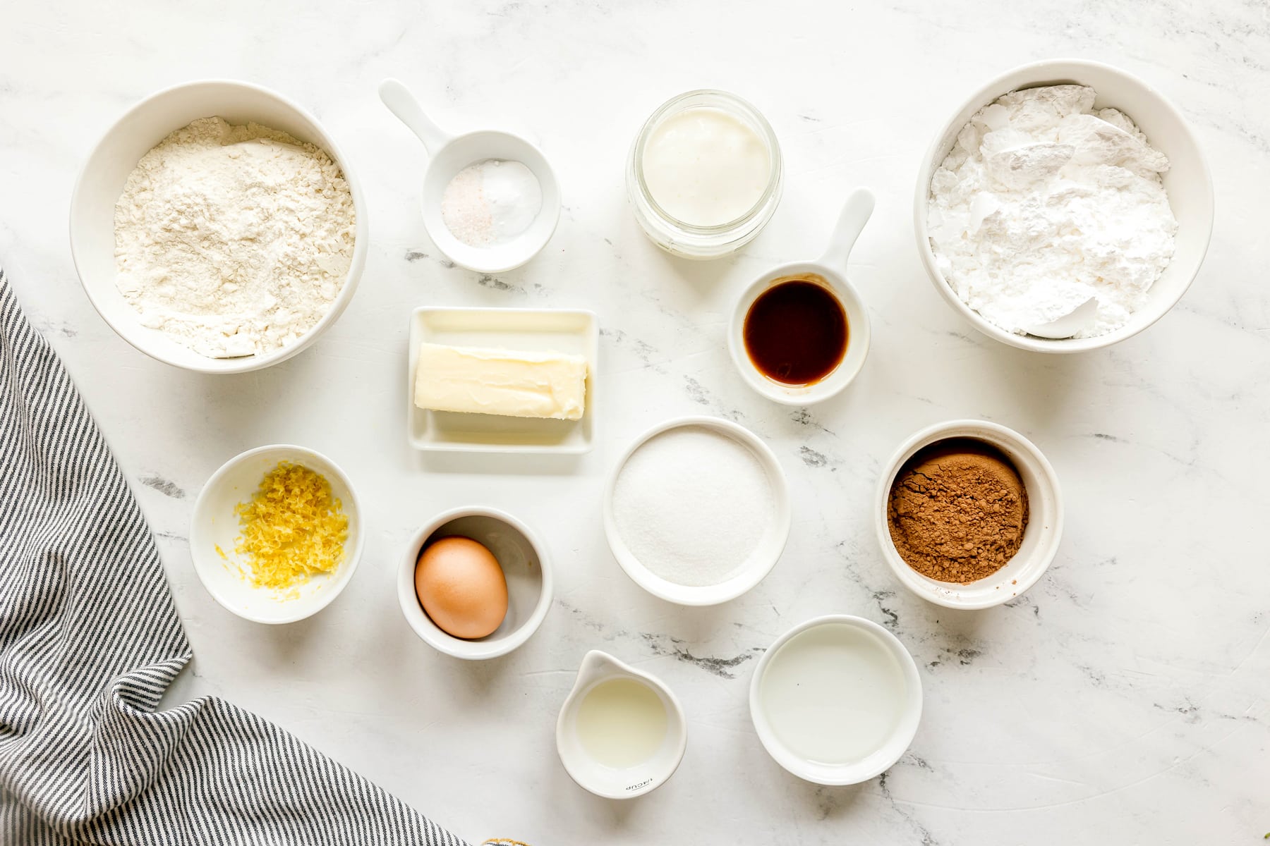 Various baking ingredients for Black and White Cookies recipe includes: all-purpose flour, baking soda, salt, buttermilk, vanilla, butter, sugar, egg, lemon zest. For the icing the ingredients are confectionaire's sugar, corn syrup, vanilla, lemon juice, and cocoa powder.