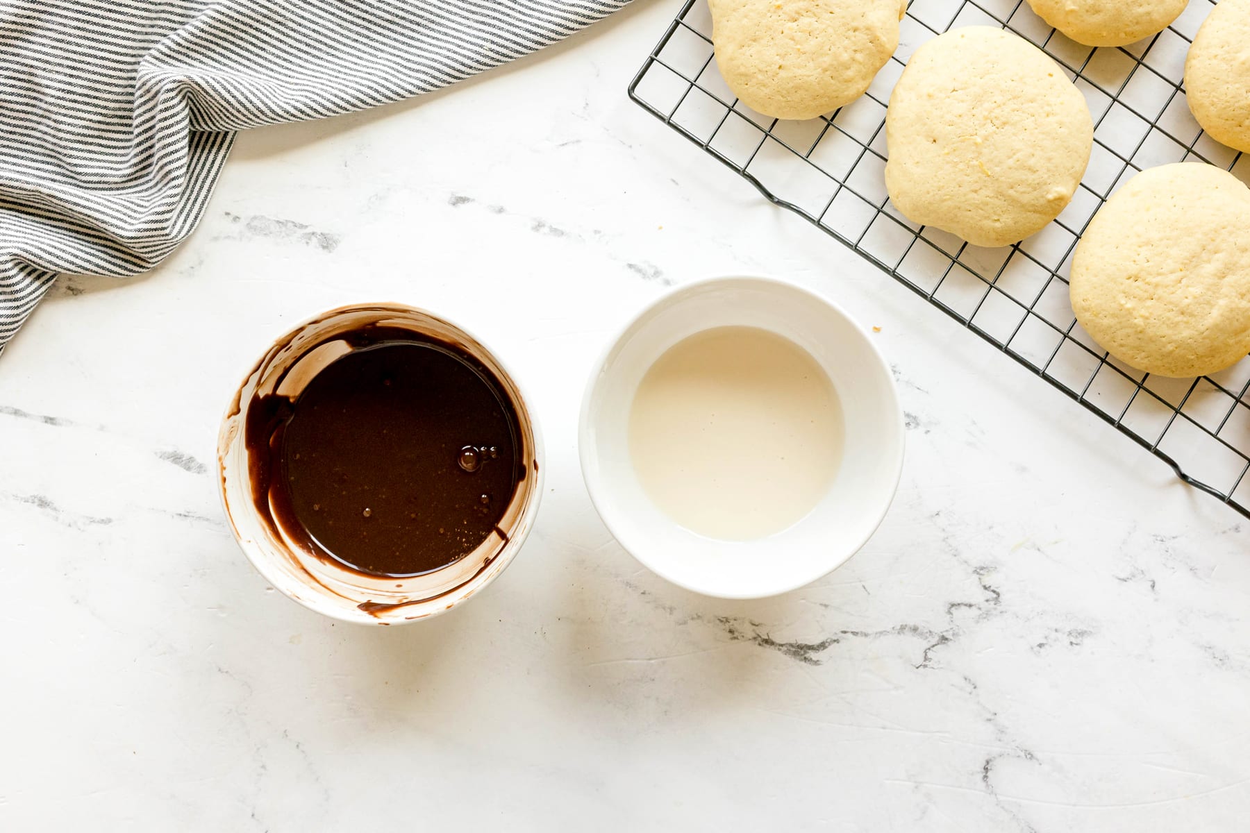 A bowl of chocolate glaze and a bowl of white glaze sit on a marble surface for Black and White Cookies recipe, next to round cookies cooling on a wire rack.