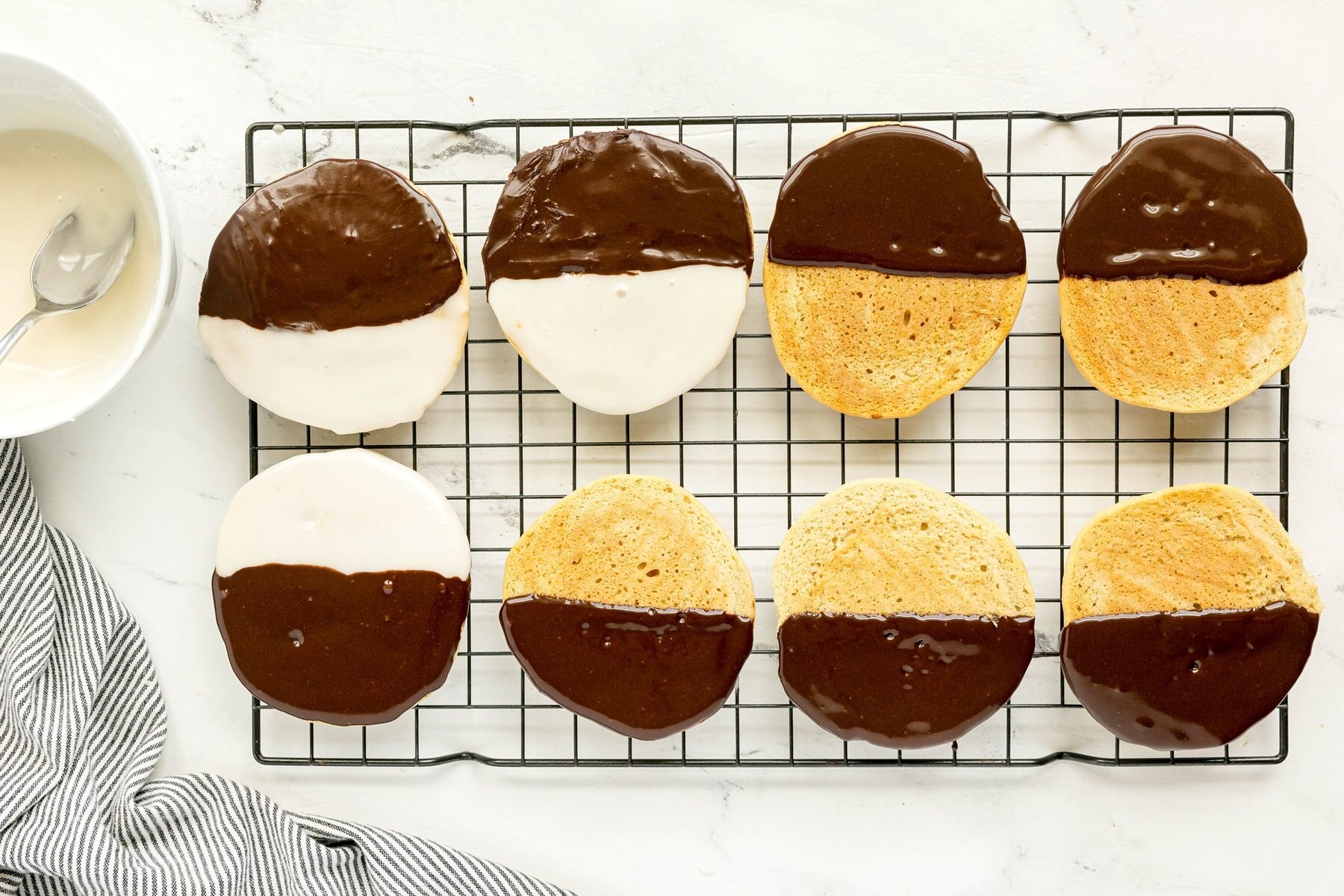 Eight Black and White Cookies cooling on a wire rack, some with icing and some plain, with a bowl and spoon in the background.