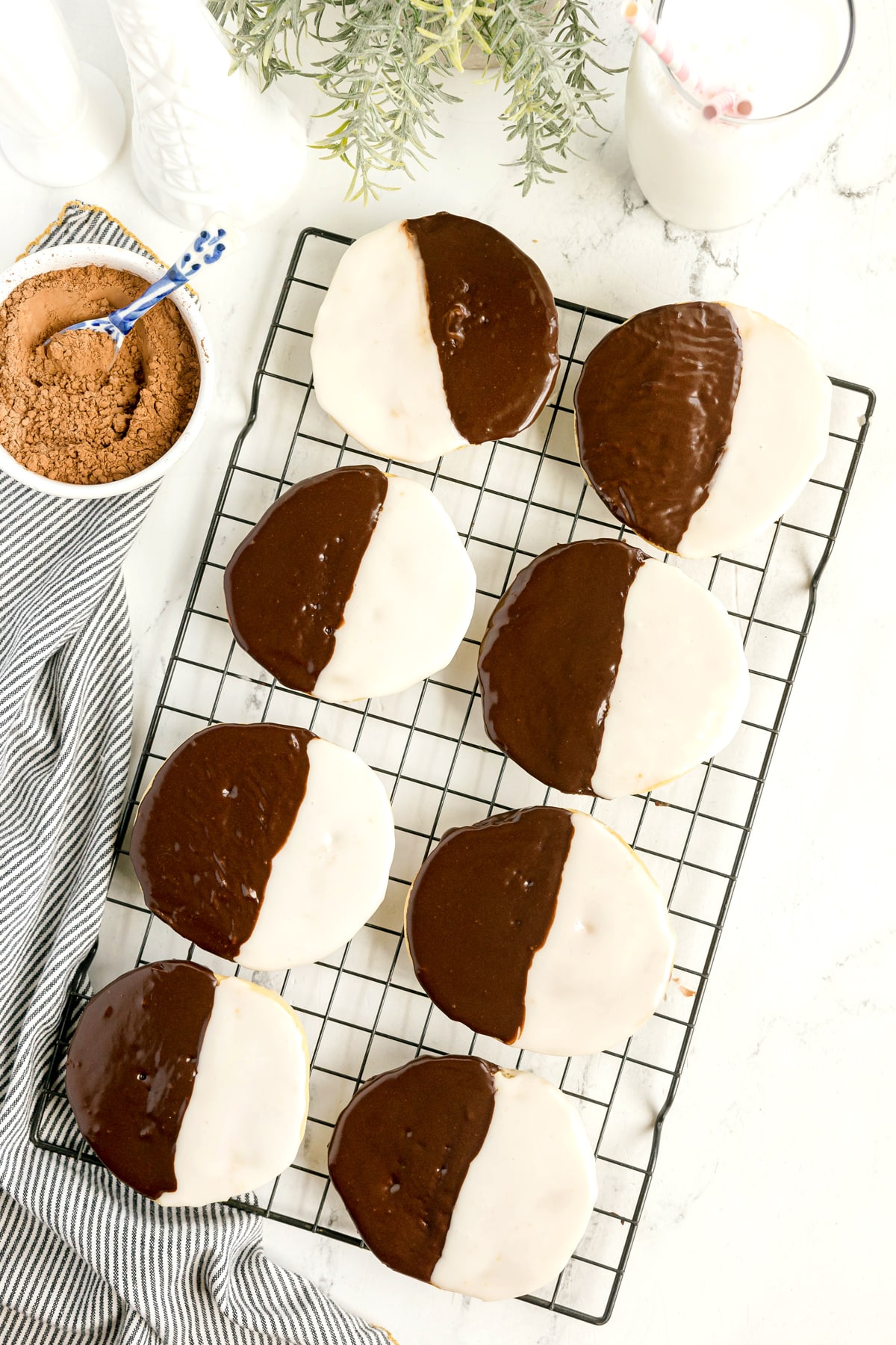 Nine Black and White Cookies are cooling on a wire rack, next to a bowl of cocoa powder, a glass of milk with a straw, and striped cloth on a white countertop.