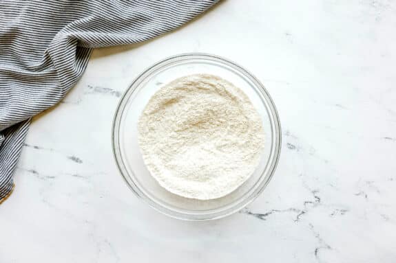 First step in preparing Black and White Cookies is to use a glass bowl filled with flour, baking soda and salt, sits on a white marble surface next to a folded, striped cloth in the upper left corner.