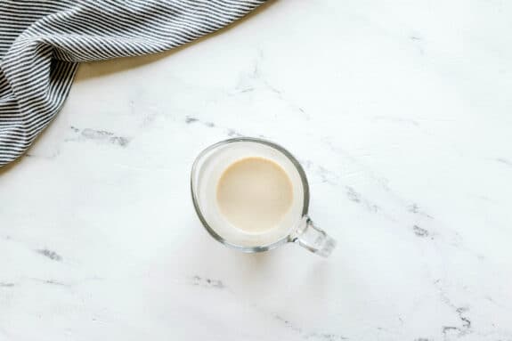 Next step in preparing Black and White Cookies is to add butter and vanilla in a cup. It sits on a white marble surface next to a black and white striped cloth.
