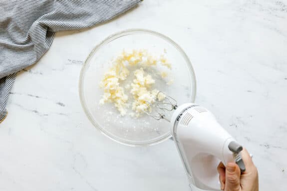 A hand holds an electric mixer blending butter and sugar for Black and White Cookies recipe in a glass bowl on a white countertop, with a striped cloth nearby.