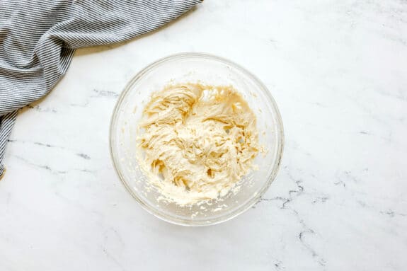 A clear glass bowl filled with creamy Black and White Cookies recipe mixture sits on a marble surface next to a striped cloth.