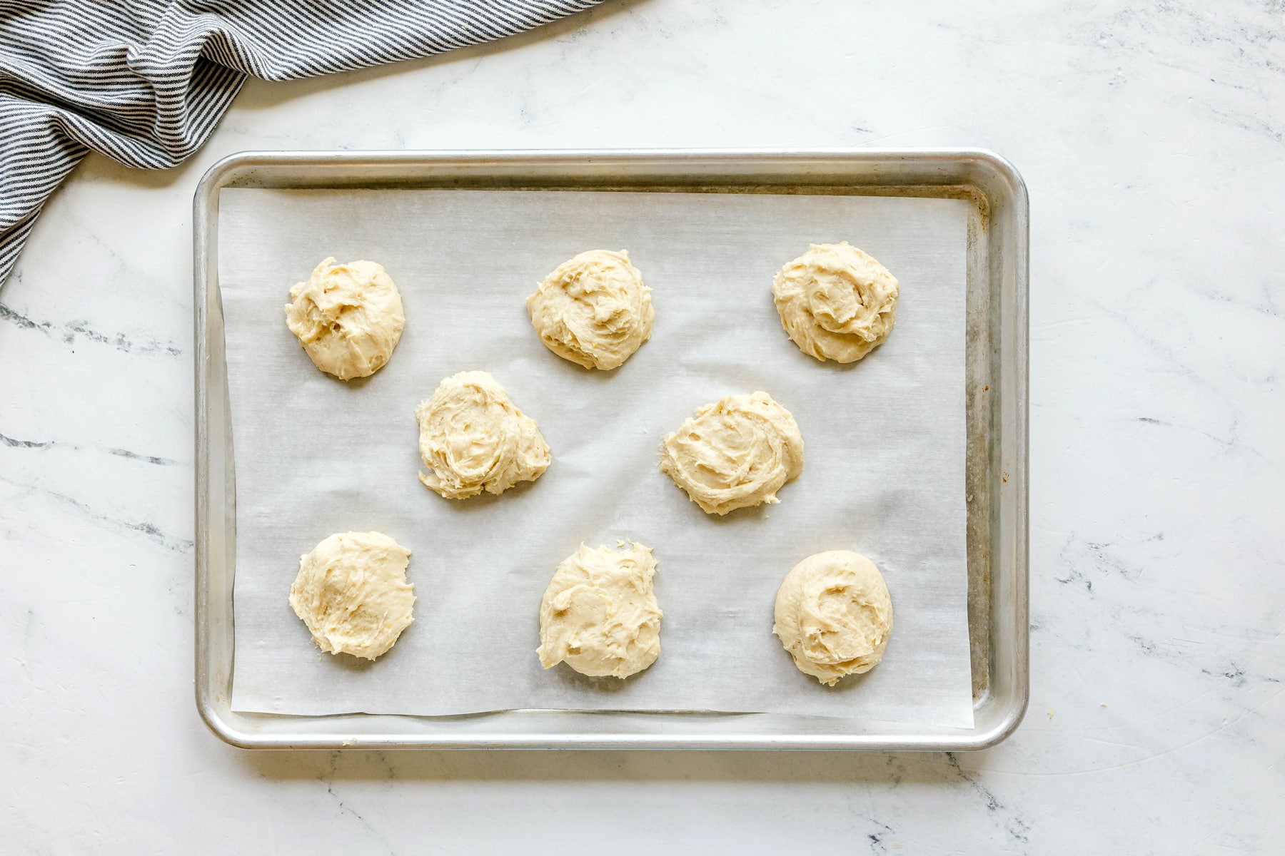 Eight unbaked Black and White Cookie dough portions on a parchment-lined baking sheet, placed on a white marble surface with a striped cloth nearby.