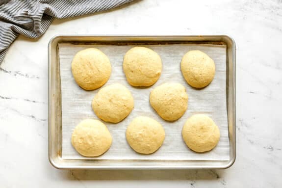 Eight round, golden cookies sit on a parchment-lined baking sheet for Black and White Cookies, atop a white marble surface with a striped cloth in the corner.