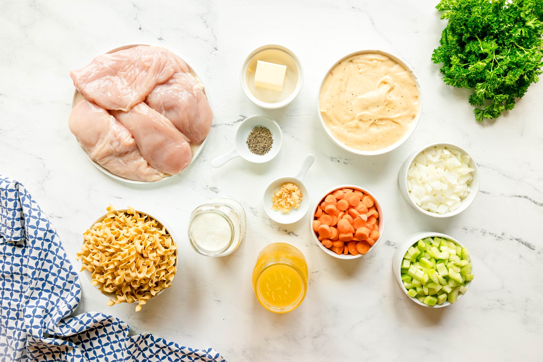 Ingredients for Slow Cooker Chicken and Noodles on a white surface, include: chicken breasts, butter, condensed cream of chicken soup, chicken stock, heavy cream, black pepper, onion, carrots, celery, minced garlic, egg noodles, and chopped parsley