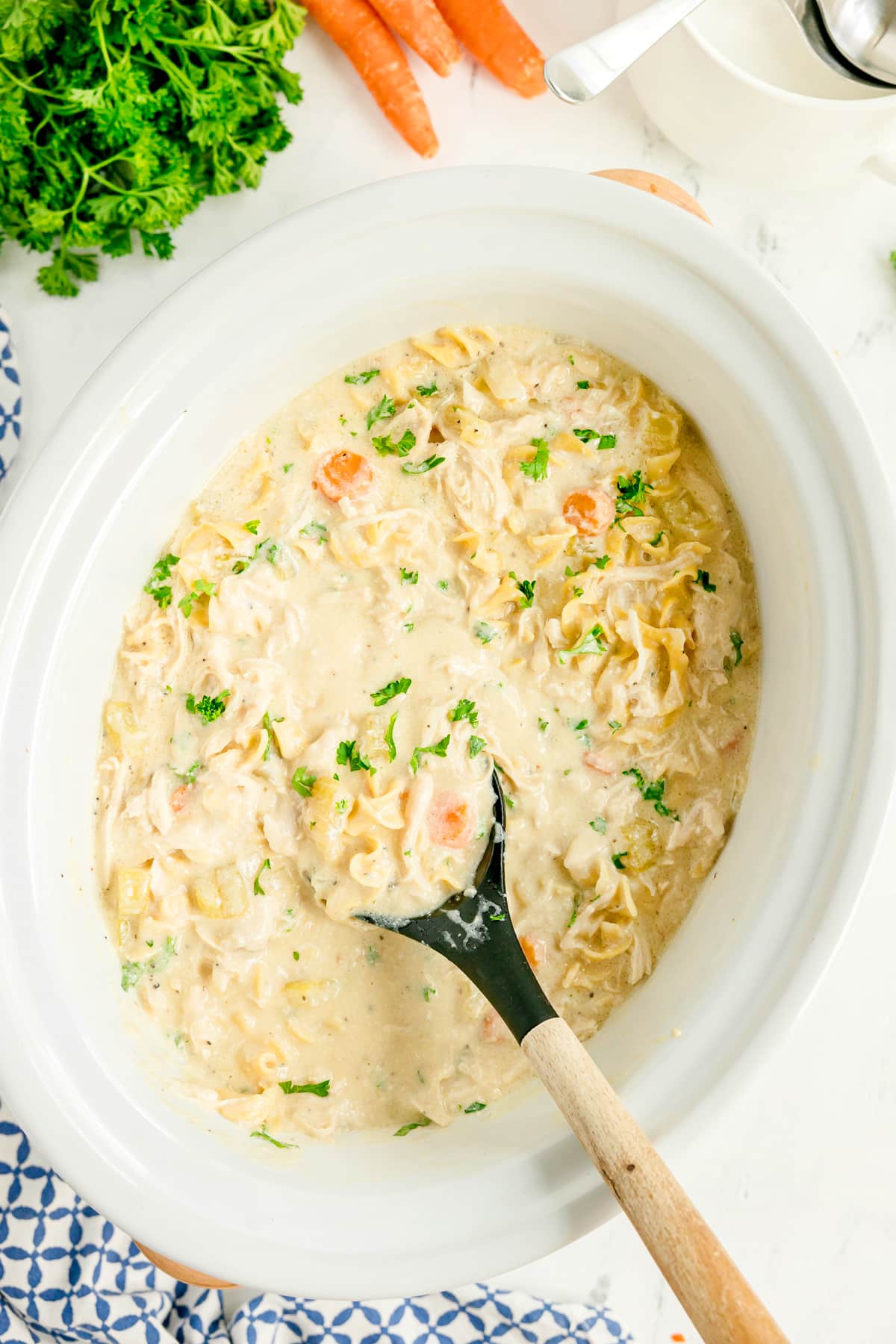 Creamy Slow Cooker Chicken and Noodles with carrots and parsley is being stirred in a white slow cooker with a black ladle.