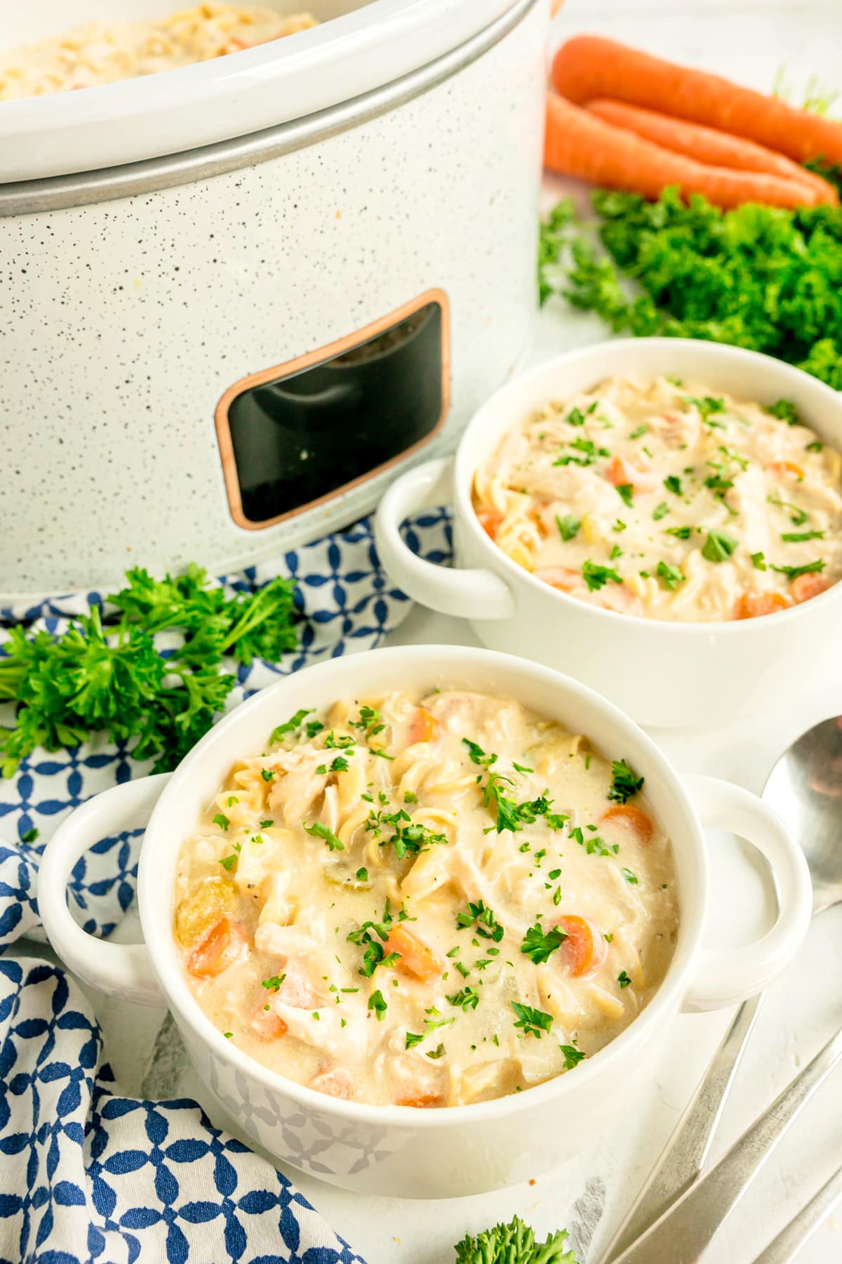 Two bowls of Slow Cooker Chicken and Noodles garnished with parsley, next to a slow cooker, fresh carrots, and parsley on a blue patterned cloth.