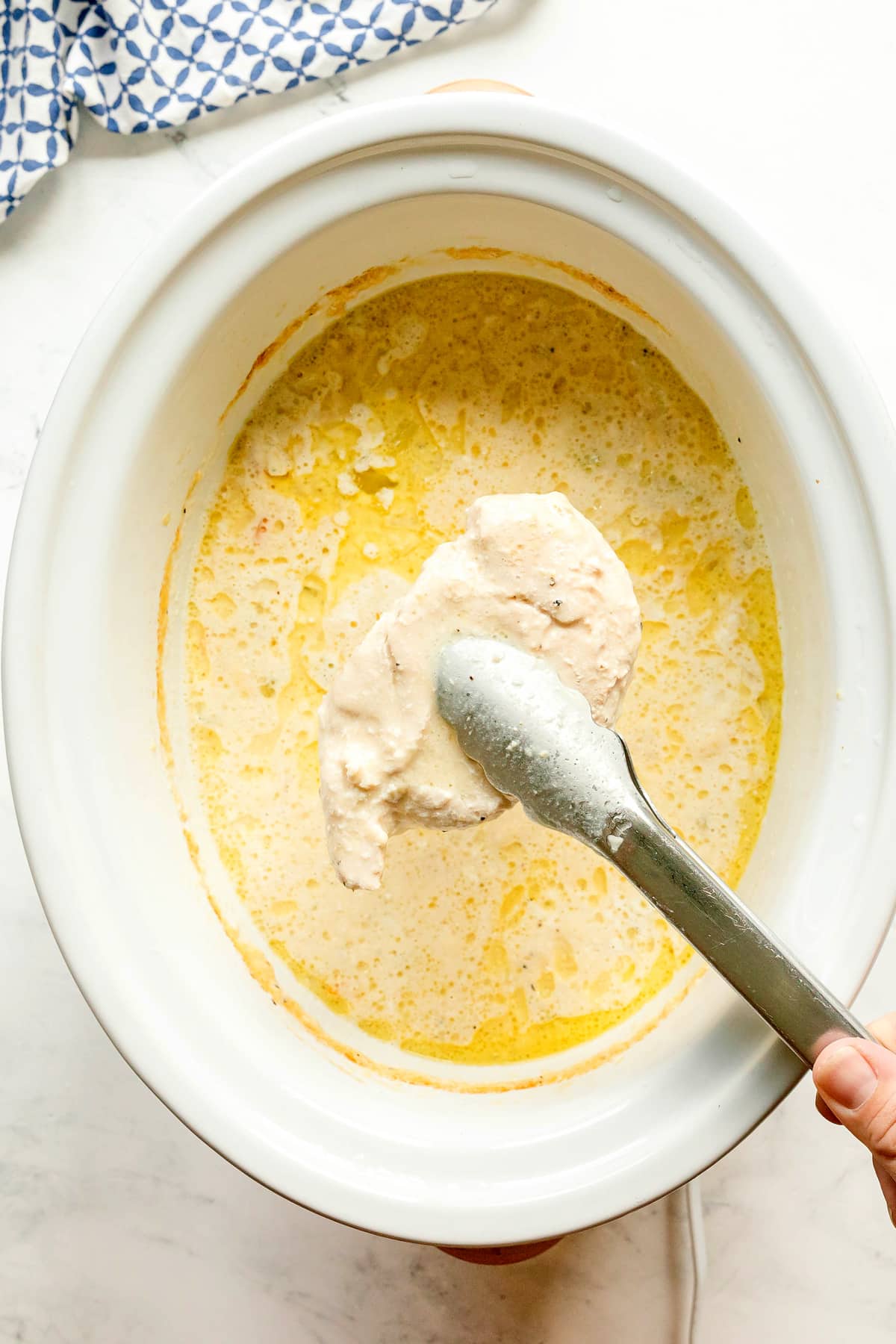 A pair of tongs removing a creamy chicken over a slow cooker filled with a yellow, creamy sauce. This is one step in preparing Slow Cooker Chicken and Noodles recipe.