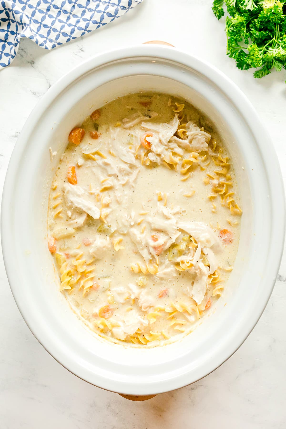 Next step in preparing Slow Cooker Chicken and Noodles is to add shredded chicken again and then add noodles, and vegetables in a creamy broth inside a white slow cooker on a light surface.