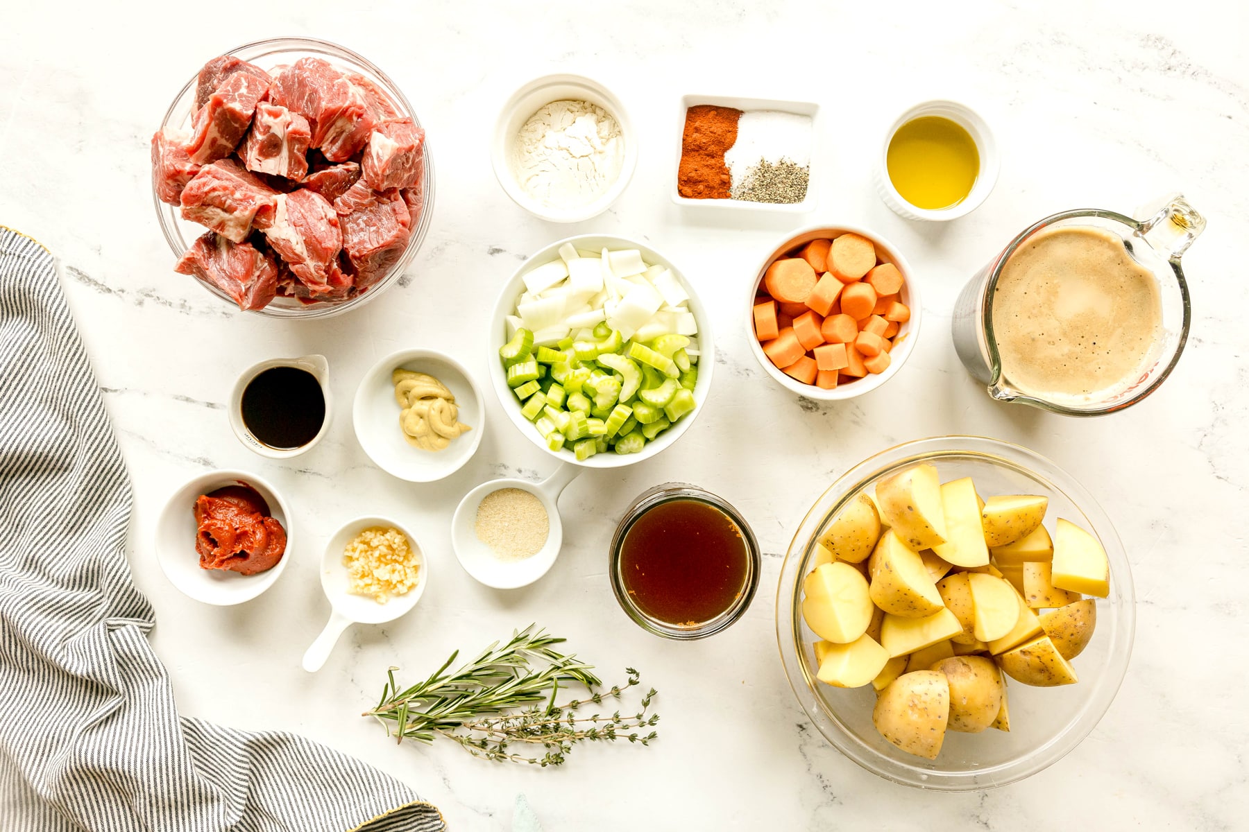A countertop with bowls of ingredients for Crockpot Guinness Beef Stew are the following: chuck roast, flour, salt, black pepper, paprika, olive oil, Guinness extra stout, large carrots, onion, celery, garlic, yukon gold potatoes, beef stock, tomatoe paste, worcestershire sauce, thyme, rosemary, onion powder, dijon mustard, and chopped parsley, arranged neatly for recipe preparation.