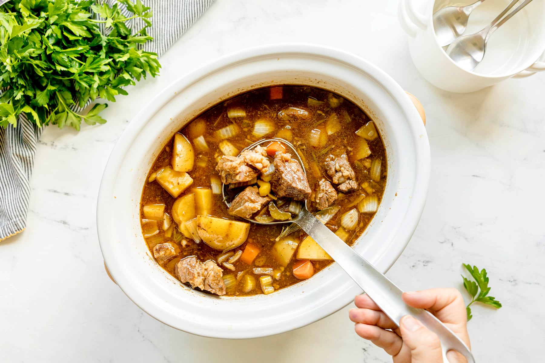 A hand holds a ladle of Crockpot Guinness Beef Stew with potatoes and carrots over a white slow cooker, with parsley and utensils nearby on a marble surface.