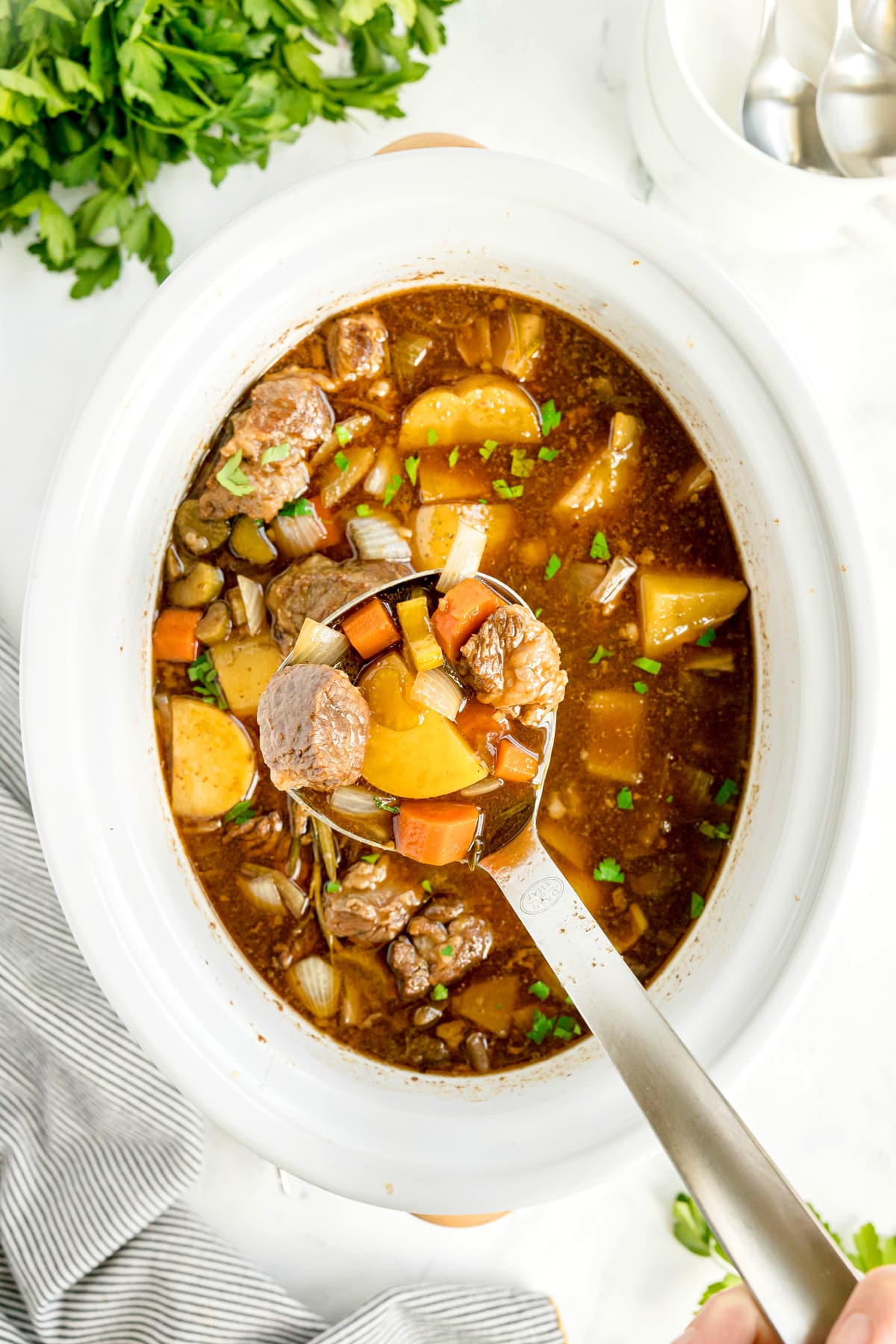 A ladle lifting Crockpot Guinness Beef Stew with potatoes, carrots, and onions from a white slow cooker; chopped parsley and utensils nearby.