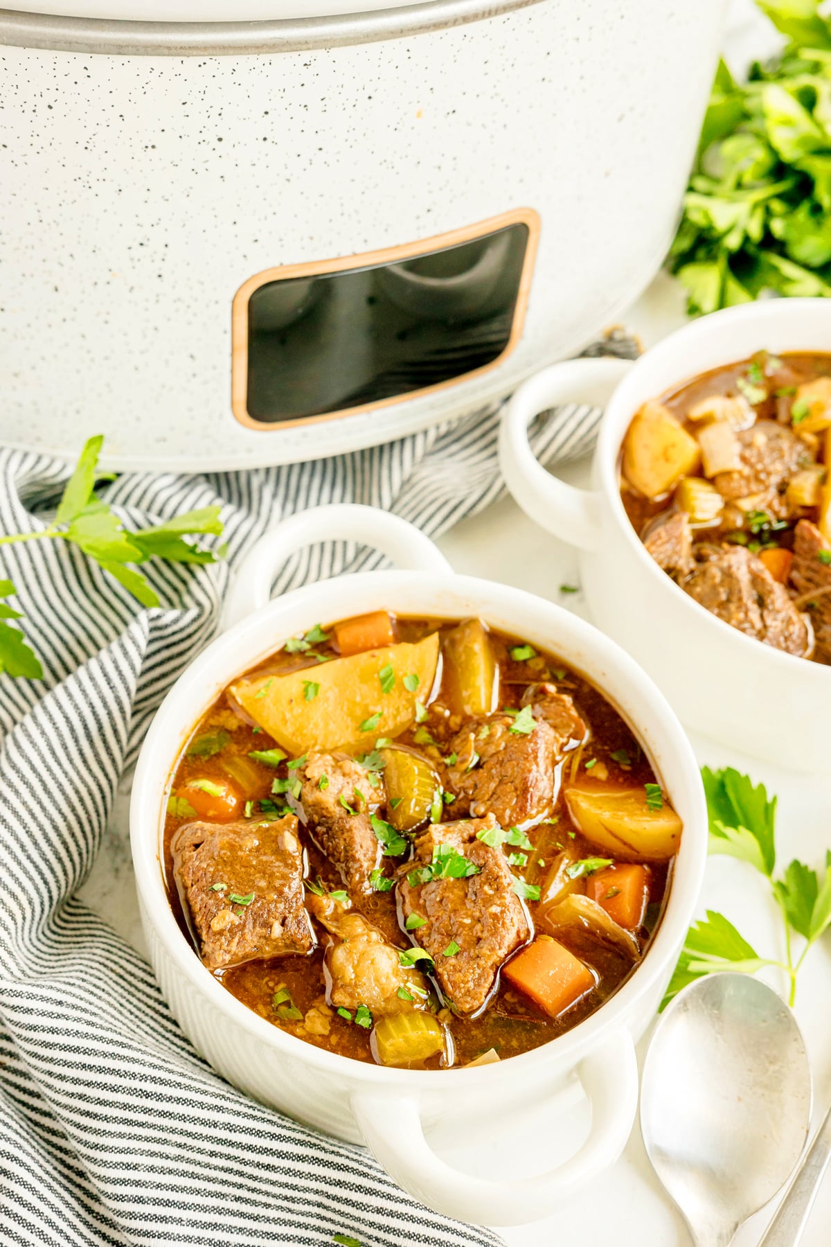 Two bowls of Crockpot Guinness Beef Stew with carrots, potatoes, and herbs sit beside a slow cooker, surrounded by parsley and a striped napkin.