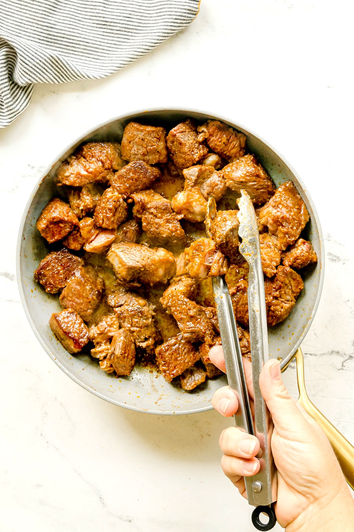 A hand uses metal tongs to pick up a piece of cooked beef for Crockpot Guinness Beef Stew ,from a skillet filled with seared beef chunks on a light countertop.