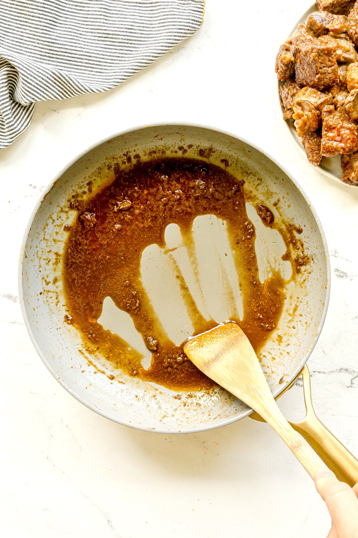 Next step in preparing Crockpot Guinness Beef Stew is to deglaze the pan with a splash of Guinness. Using a wooden spatula scrape browned bits and sauce from a skillet. Cooked meat pieces on a plate can be seen nearby.