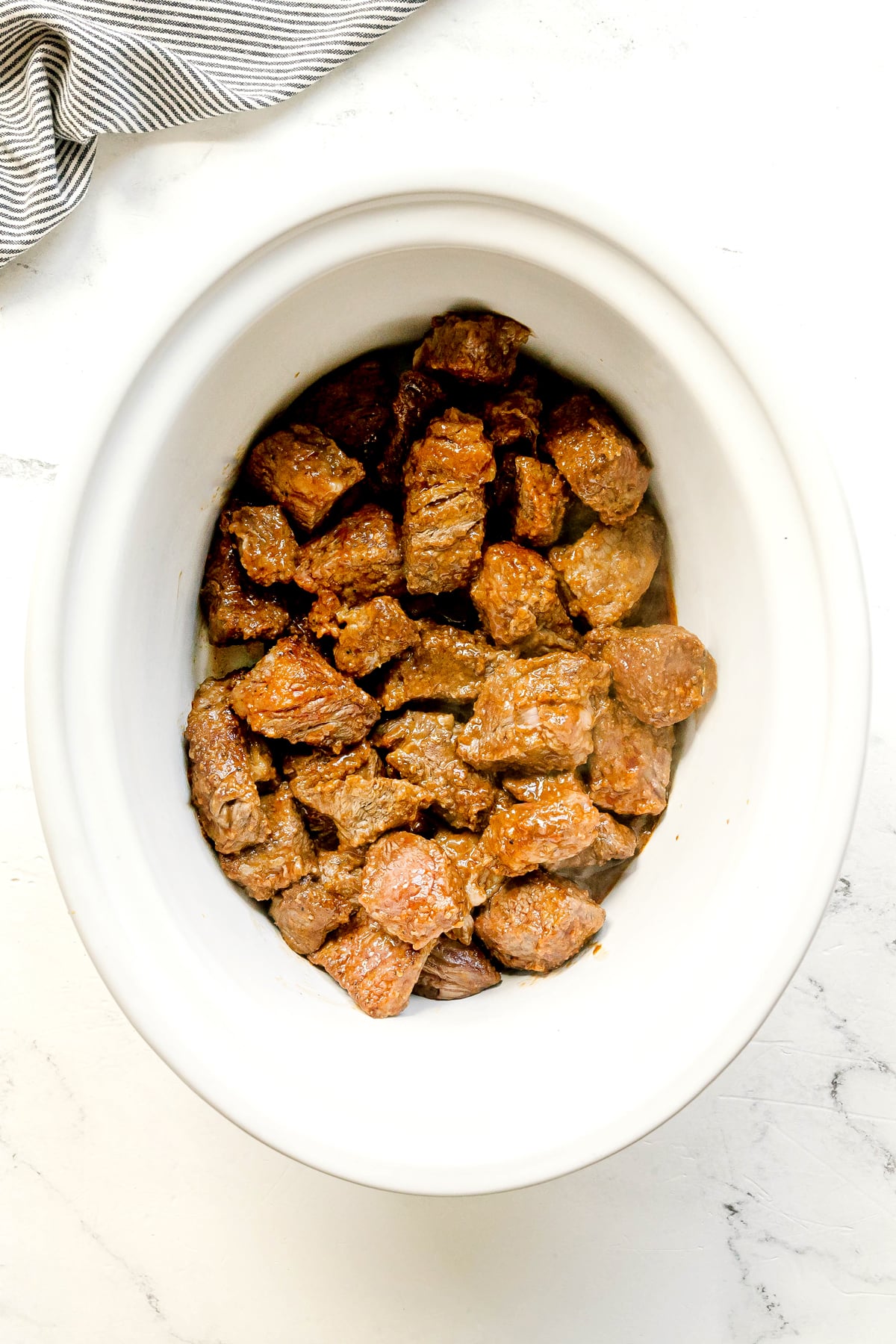 Then the next step in preparing Crockpot Guinness Beef Stew is to add chunks of browned beef in a crockpot on a light countertop, with a striped cloth partially visible in the top left corner.