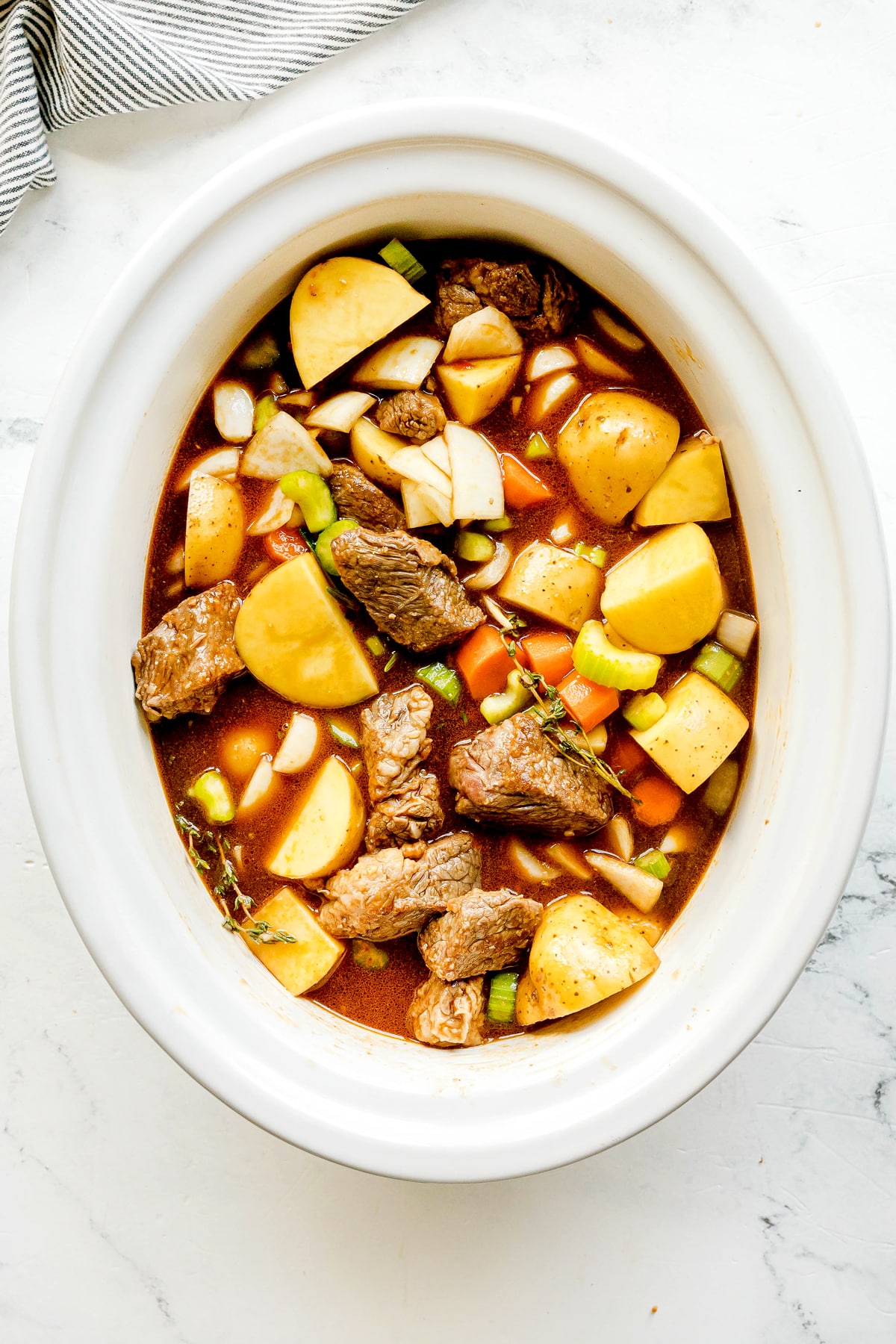 A white crockpot filled with raw beef chunks, potato pieces, carrots, celery, onion, broth, and herbs, ready to be cooked. This is Crockpot Guinness Beef Stew recipe in the making.
