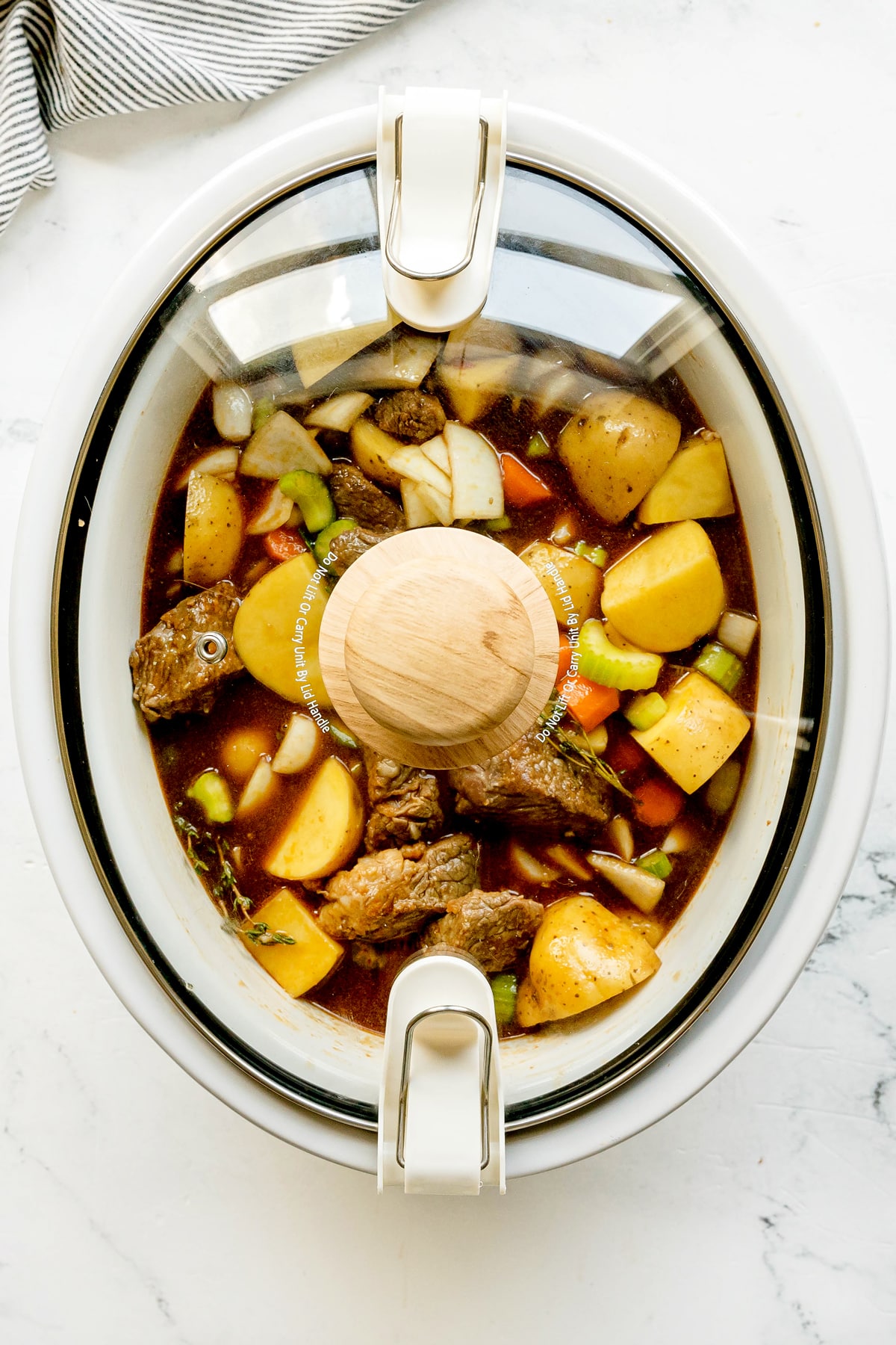 Slow cooker filled with Crockpot Guinness Beef Stew ingredients , including chunks of beef, potatoes, carrots, celery, and onions in broth, on a white countertop.