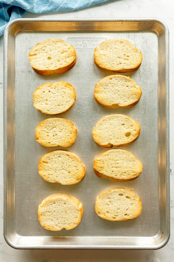 Ten slices of toasted bread are arranged in two rows on a metal baking sheet, with a blue cloth partially visible in the top left corner.