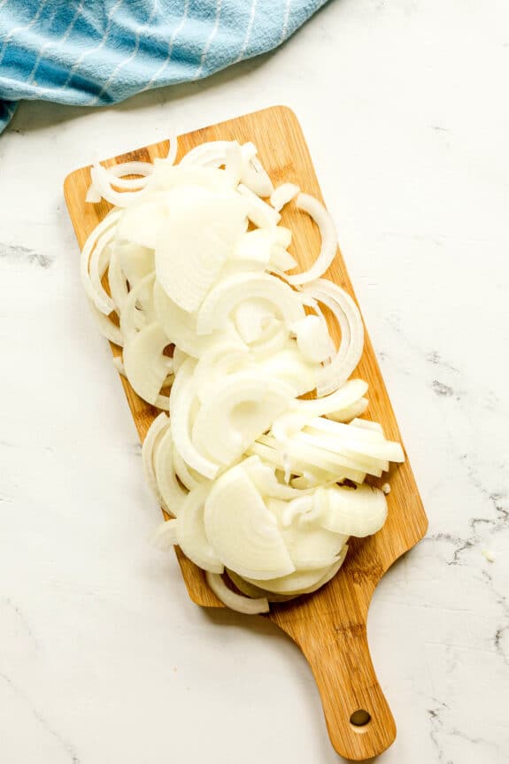 Thin slices of white onions for French Onion Soup are arranged on a wooden cutting board, set on a white surface with a blue cloth in the background.