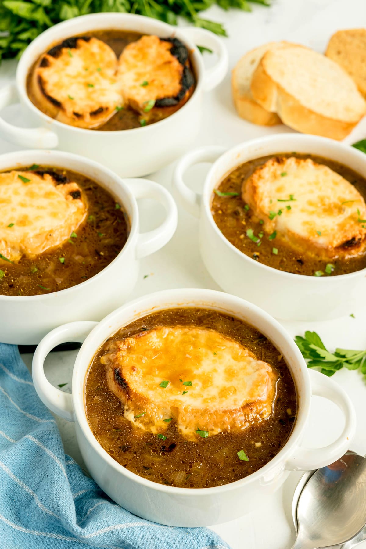 Four bowls of French onion soup topped with toasted bread and melted cheese, garnished with parsley; slices of bread and fresh herbs in the background.