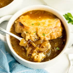 A white bowl of French onion soup with a slice of bread topped with melted cheese, a spoon lifting caramelized onions, and a blue napkin nearby.
