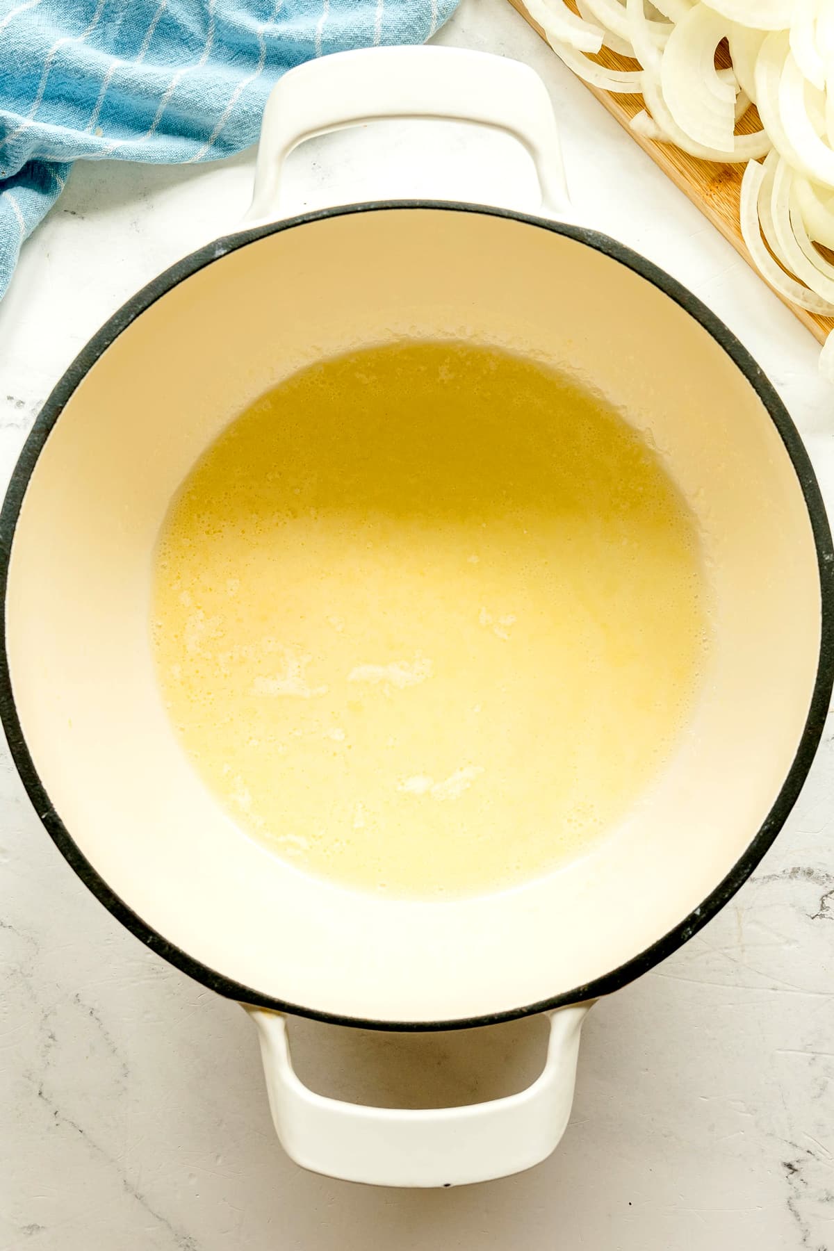 A white pot with melted butter or oil sits on a white countertop. Sliced onions and a blue towel are visible in the background. This is one of the processes in preparing French Onion Soup recipe.