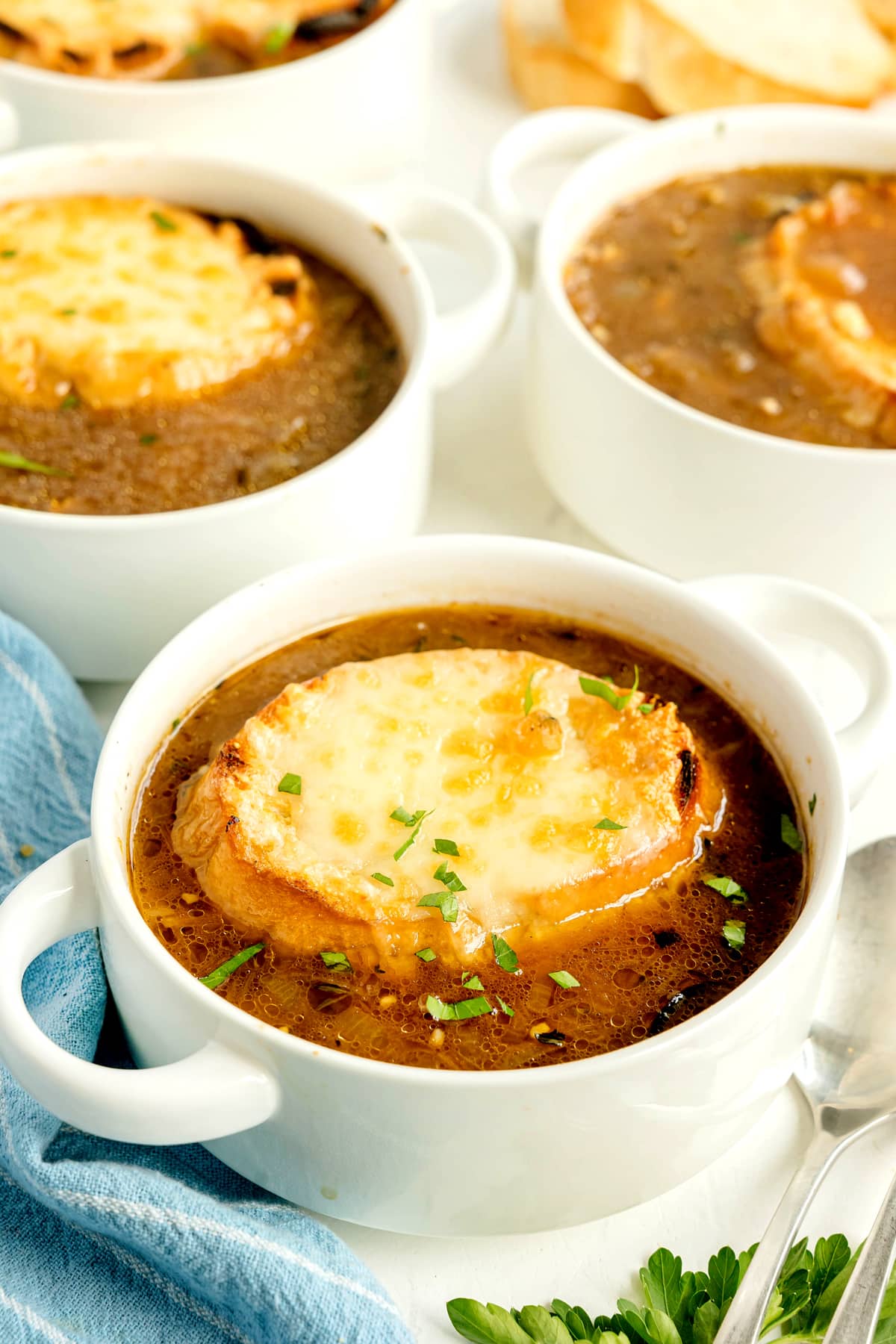 Bowls of French onion soup topped with toasted bread and melted cheese, garnished with chopped herbs, are arranged on a white surface with a spoon, napkin, and parsley nearby.
