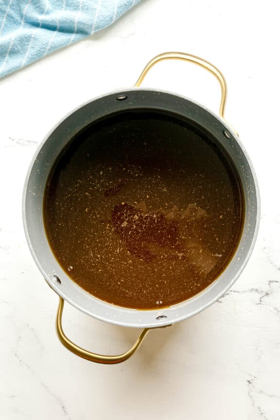 Next step in preparing French Onion Soup is to add beef stock, on a white surface with a blue-striped cloth in the corner.