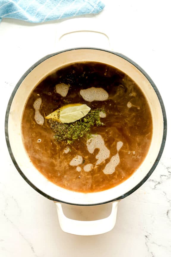 A white pot filled with brown beef broth, added with Worcestershire sauce, thyme, bay leaf, and pepper. This is the next step in preparing French Onion Soup