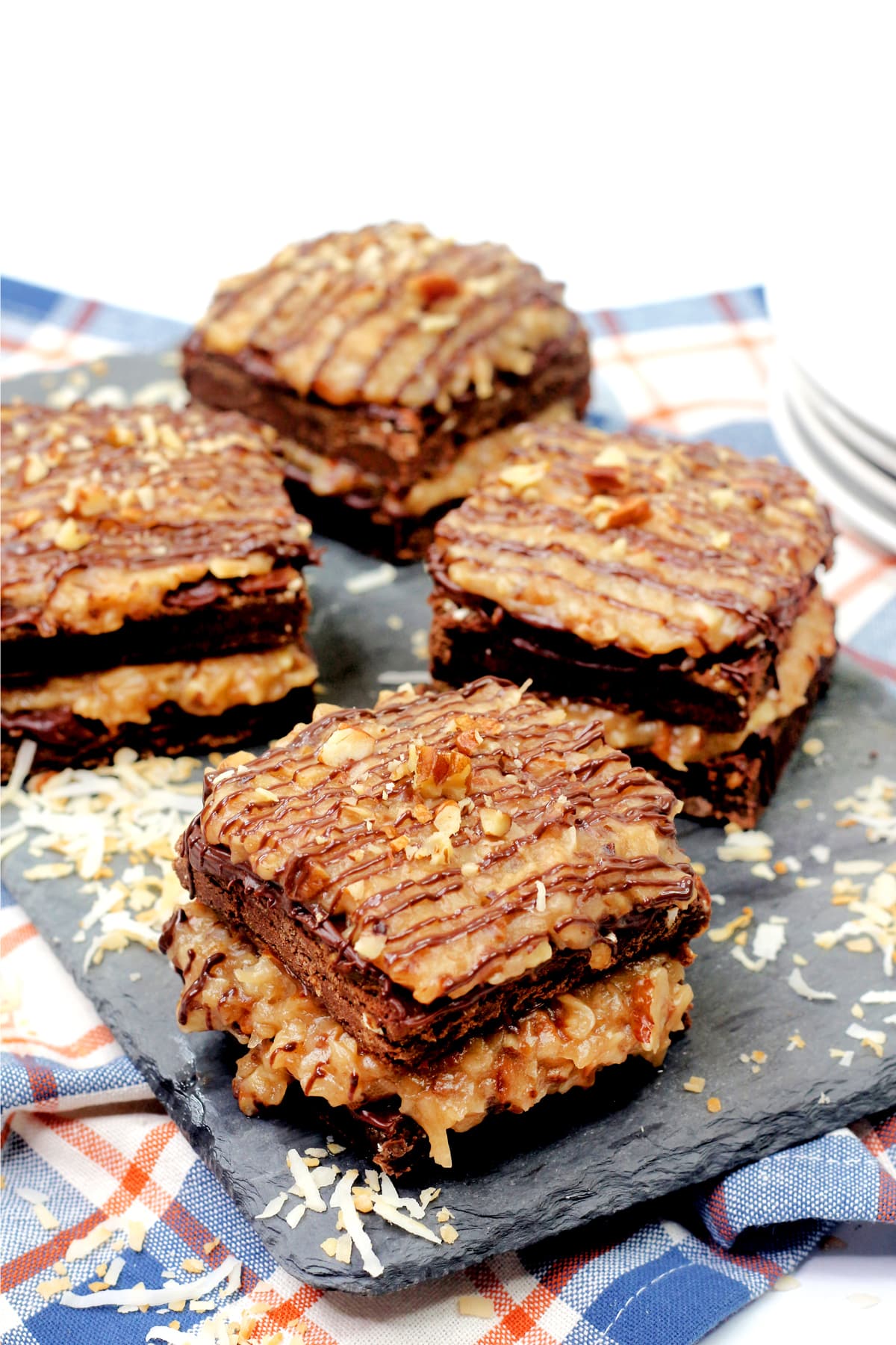 Four square German Chocolate Brownies are stacked on a slate tray, topped with chocolate drizzle, with a plaid cloth and shredded coconut in the background.