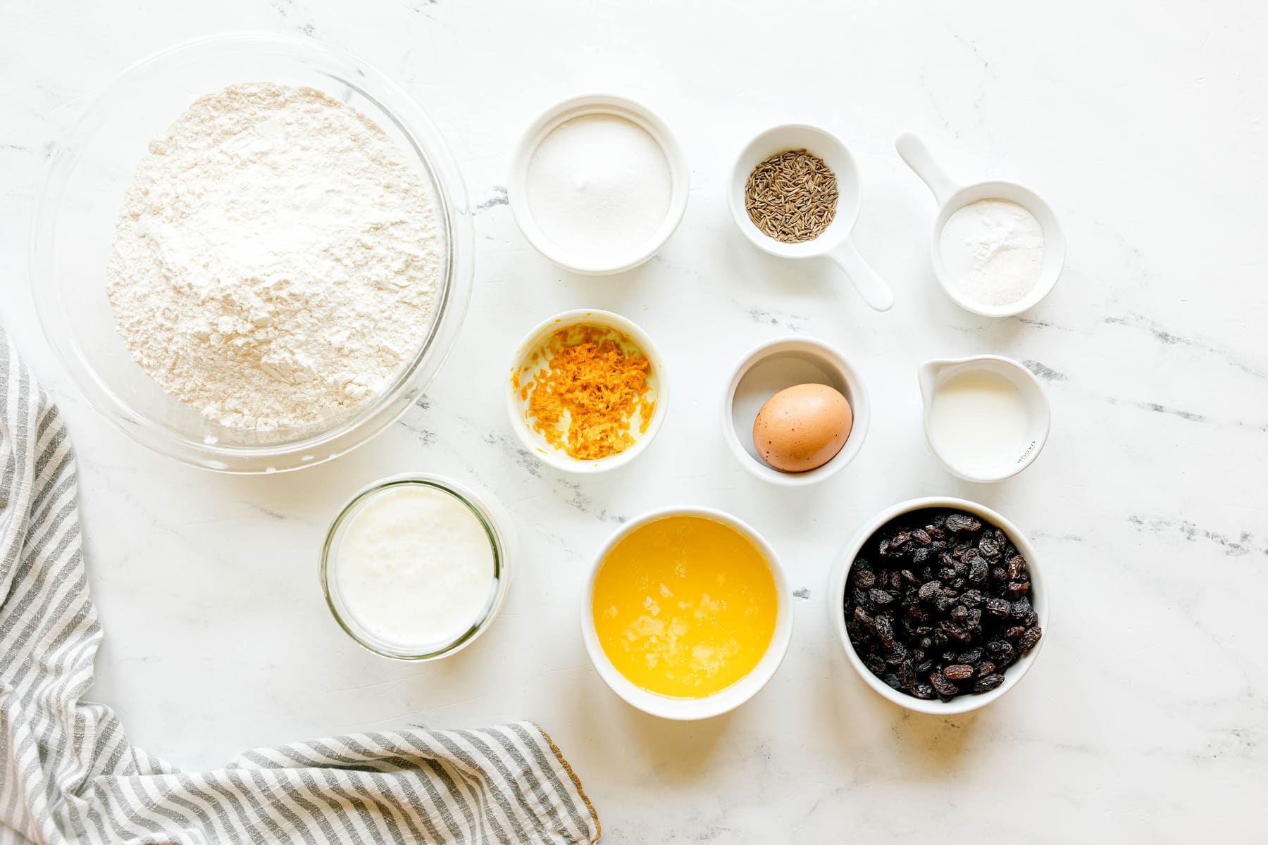 Various baking ingredients for for Irish Soda Bread in bowls and cups on a white surface, including all-purpose flour, baking soda, baking powder, salt, caraway seeds, buttermilk, sugar, butter, egg, orange zest, milk, and raisins or currants