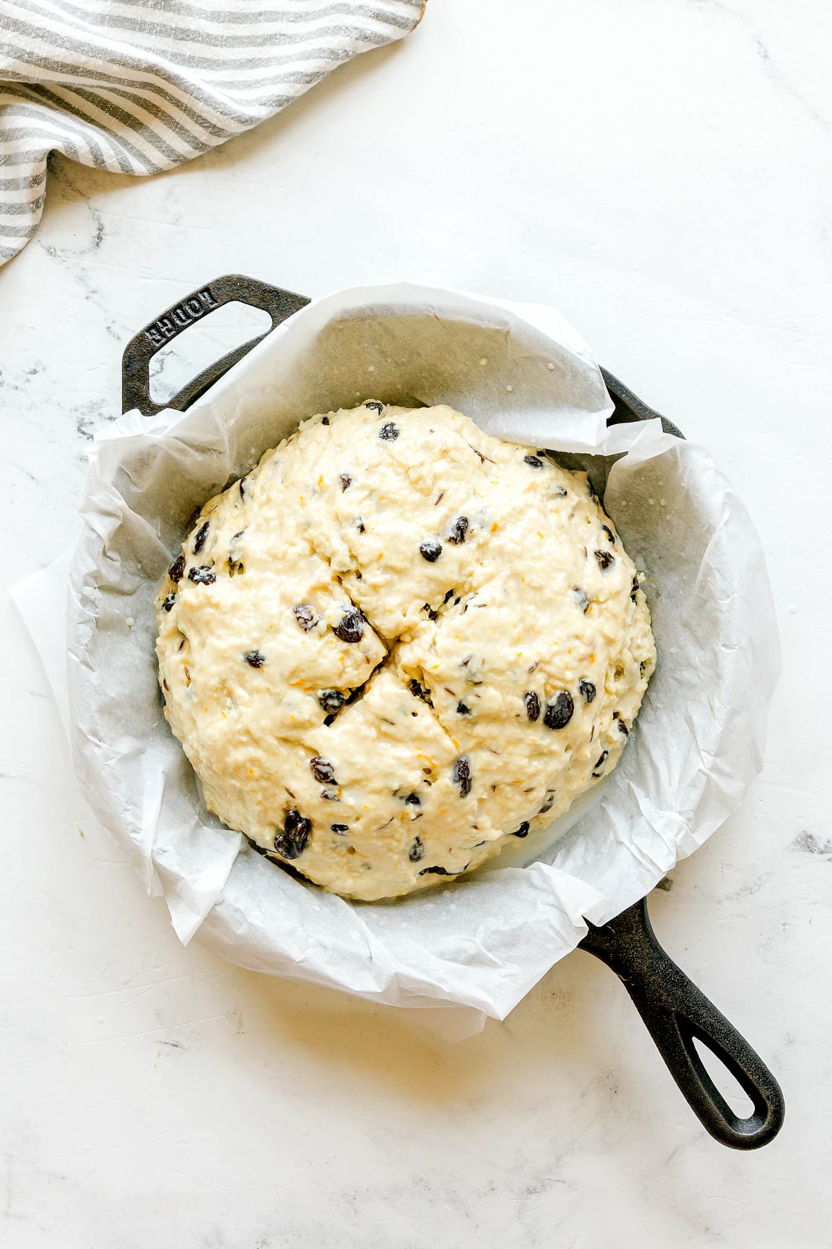Unbaked Irish Soda Bread dough with raisins sits in a parchment-lined cast iron skillet on a white surface, next to a striped cloth.