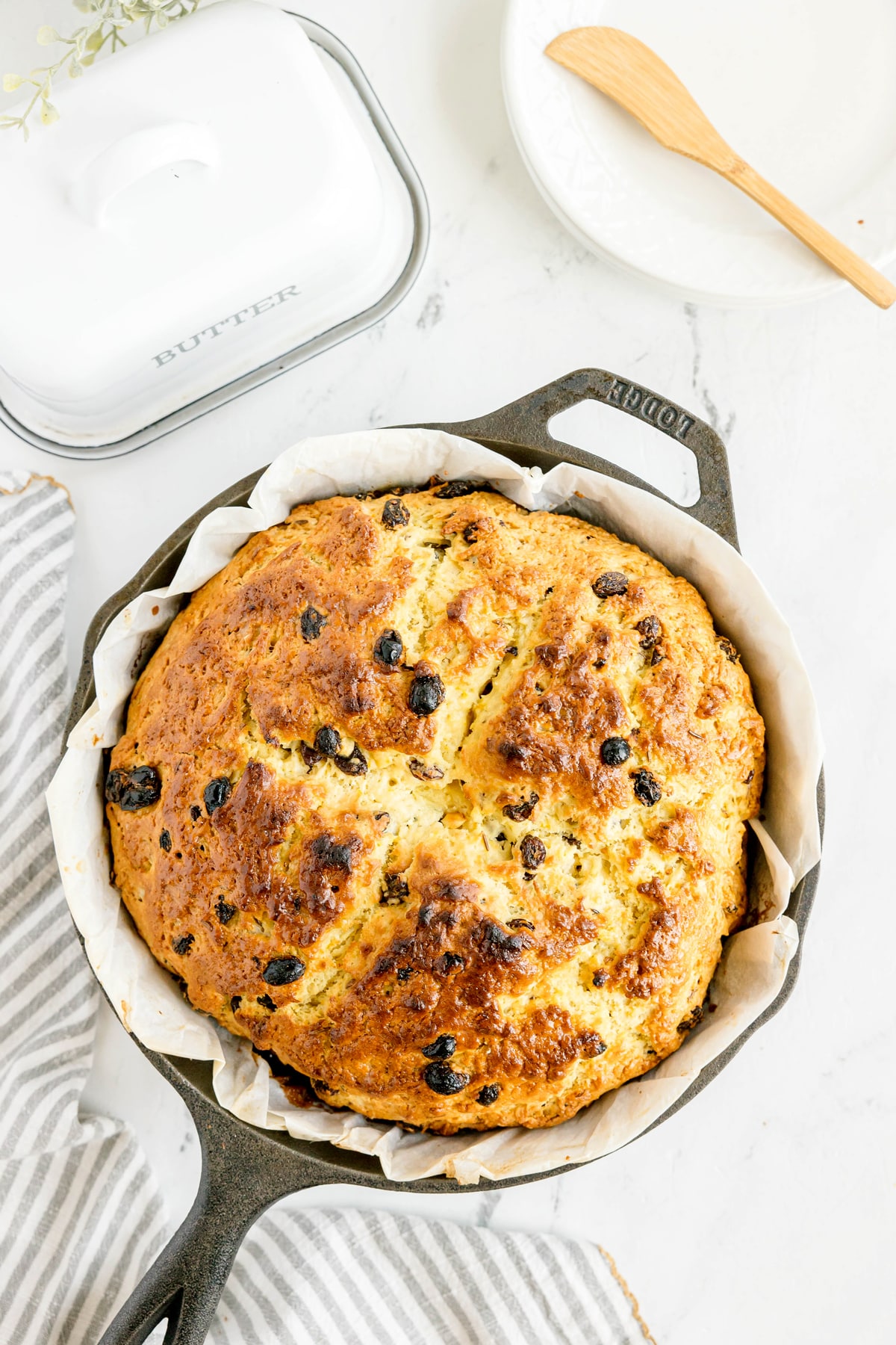 A round loaf of Irish Soda Bread with raisins in a cast iron skillet lined with parchment paper, next to a butter dish and a striped cloth.