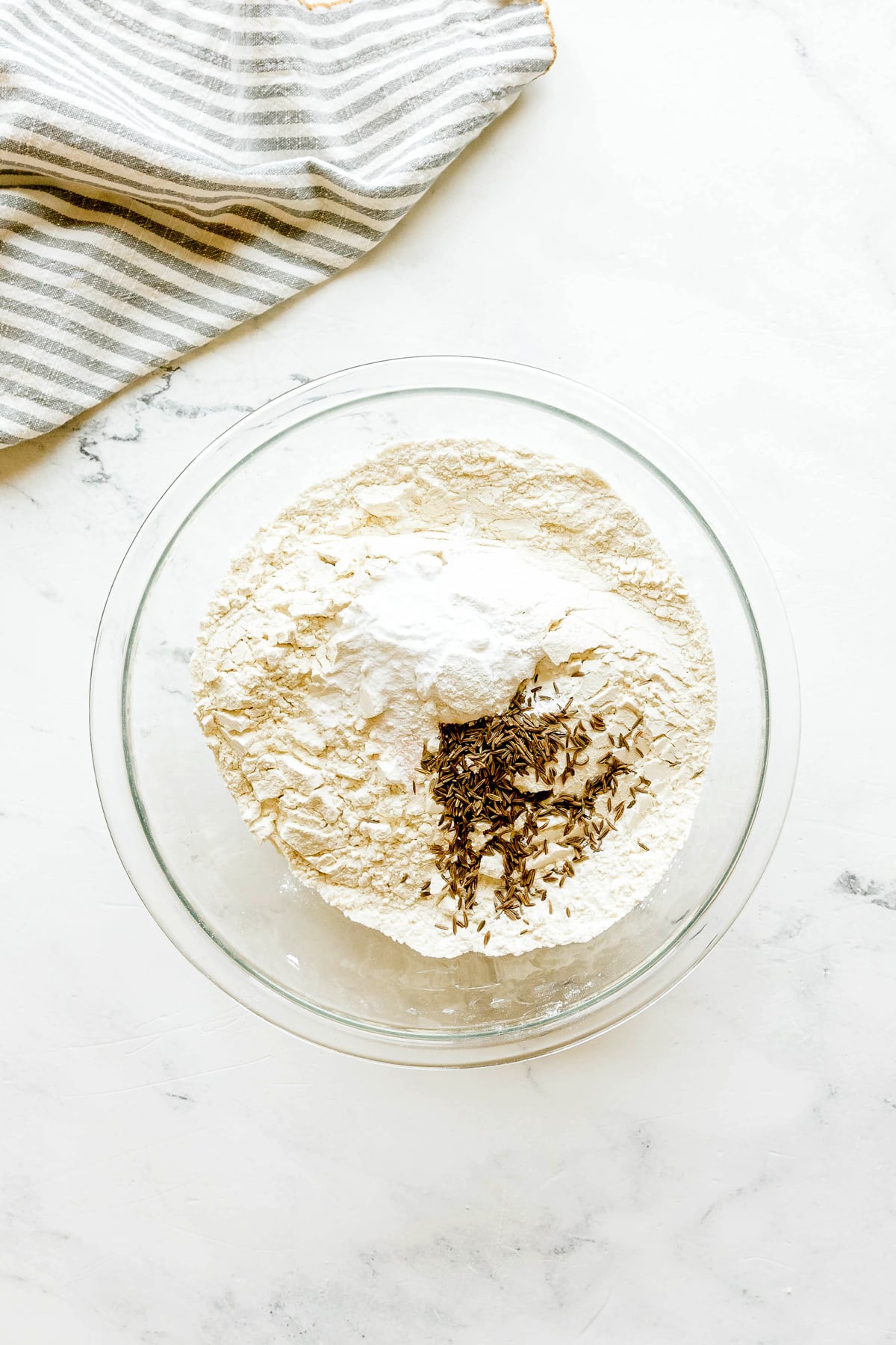 First step in preparing Irish Soda Bread is to use a glass bowl with flour, baking powder, and caraway seeds on a white surface, next to a striped cloth.