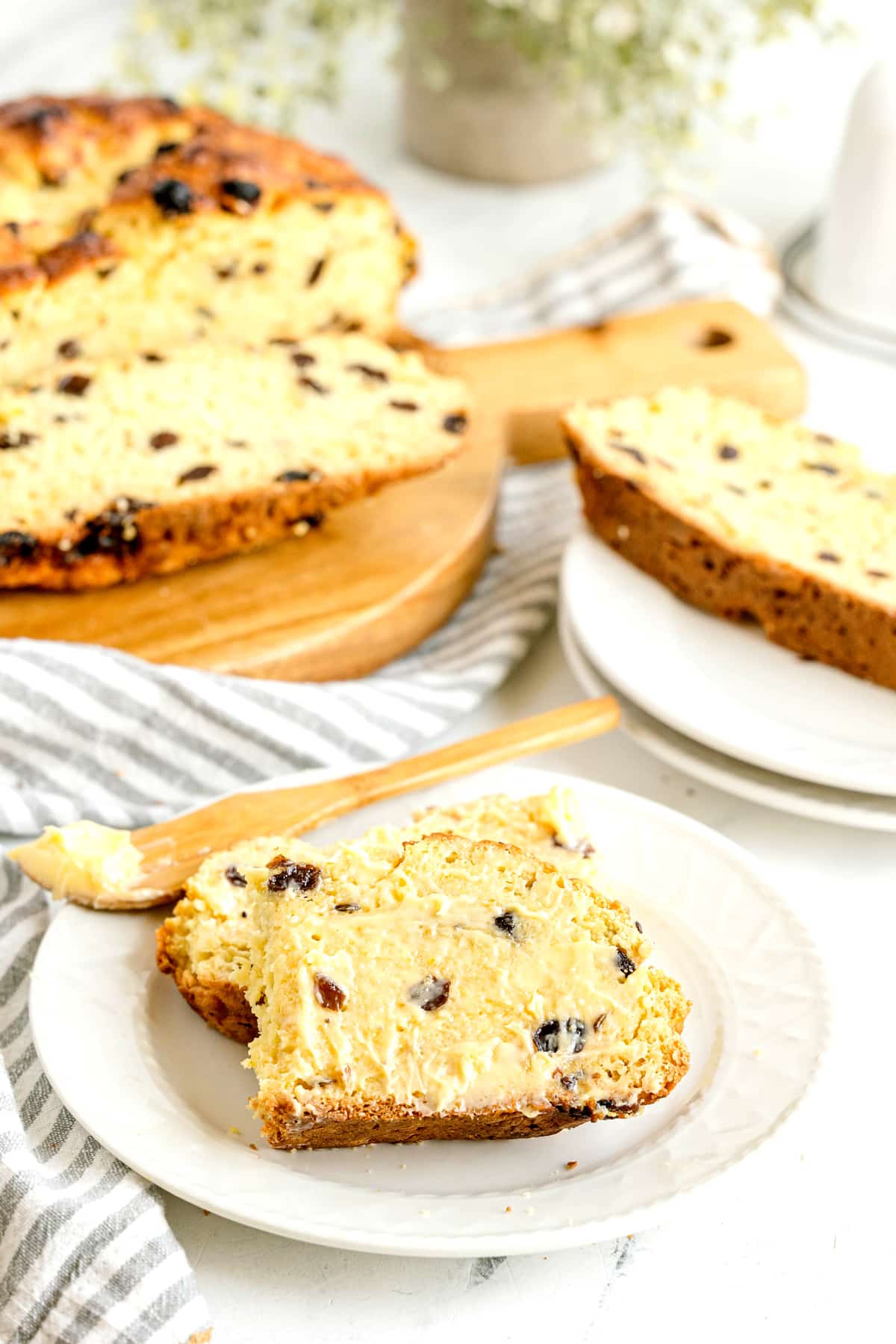 A slice of fruit-studded Irish Soda Bread spread with butter sits on a plate, with more sliced bread and a butter knife nearby on a striped napkin.