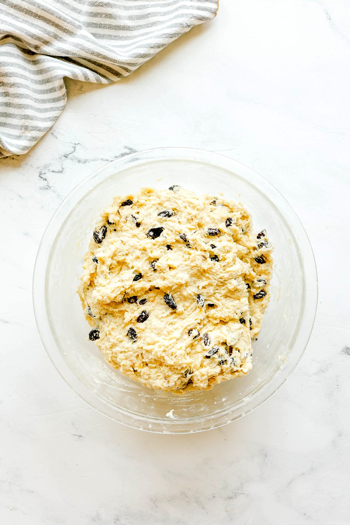 A glass bowl filled with dough mixed with raisins for Irish Soda Bread sits on a white marble surface next to a striped cloth.
