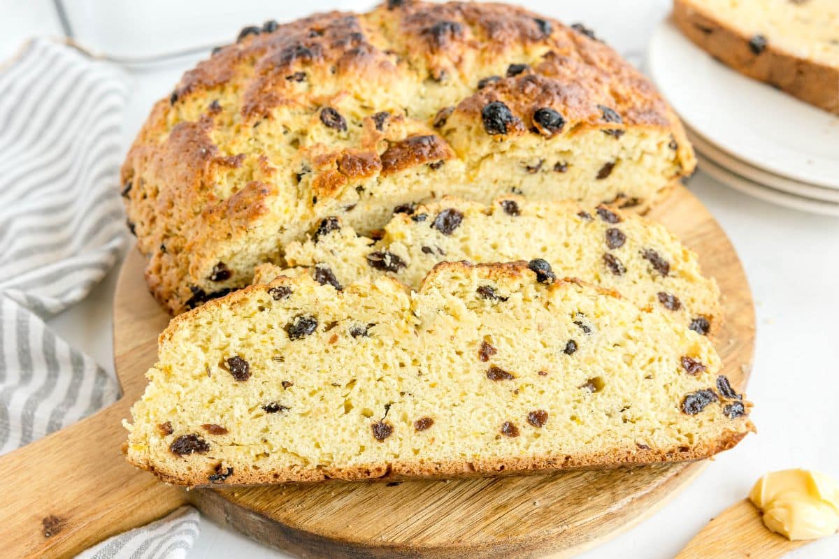 A loaf of Irish soda bread with raisins, partially sliced, sits on a wooden cutting board next to a striped towel and a stack of white plates, capturing the essence of a True Irish Soda Bread Recipe.