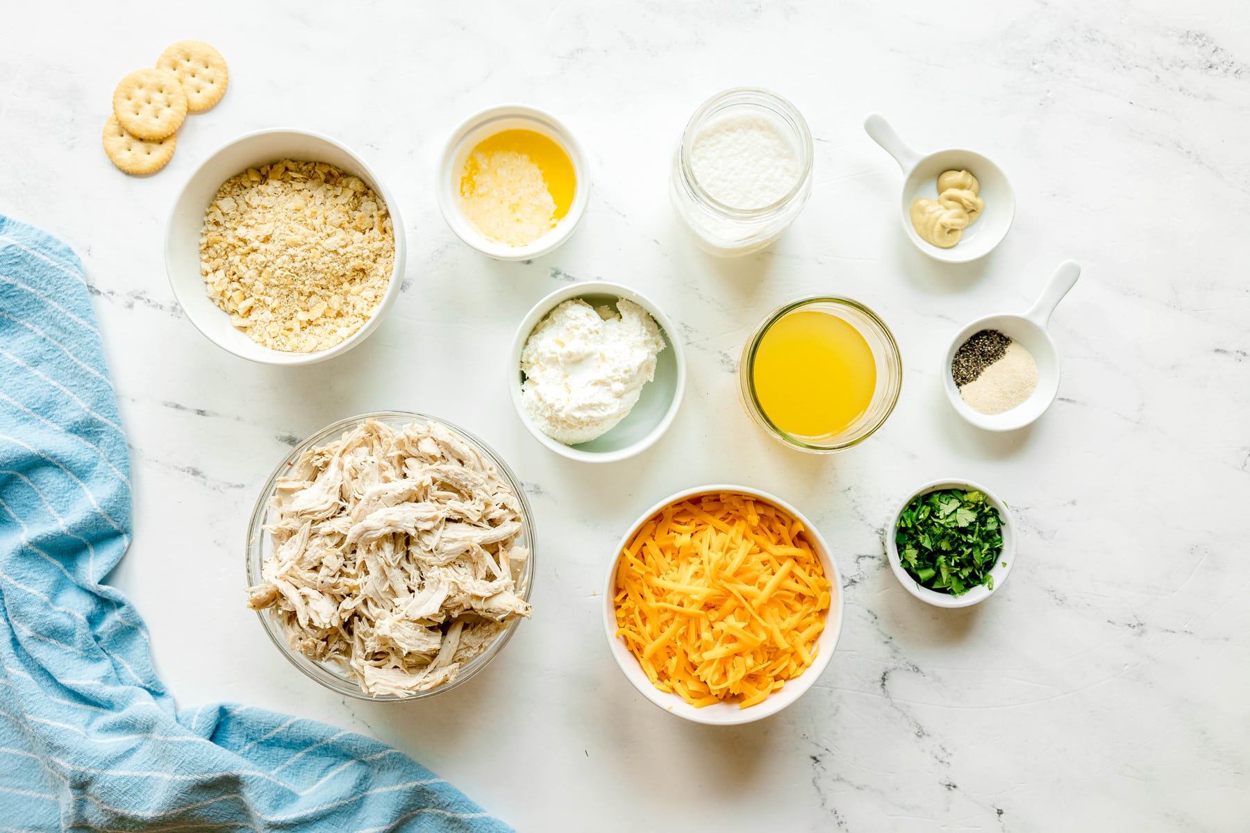 Overhead view of Ritz Cracker Casserole with 2 sleeves Ritz crackers, 3 tbsp. butter, cream cheese, heavy cream, chicken stock, Dijon mustard, onion powder, black pepper, cheddar cheese, cooked chicken and chopped parsley with a blue towel on the left.