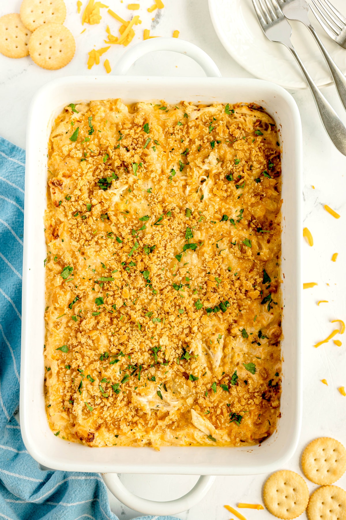 Ritz Cracker Casserole topped with golden breadcrumbs, garnished with herbs, next to a blue towel, plates, forks, shredded cheese, and round crackers on a white surface.