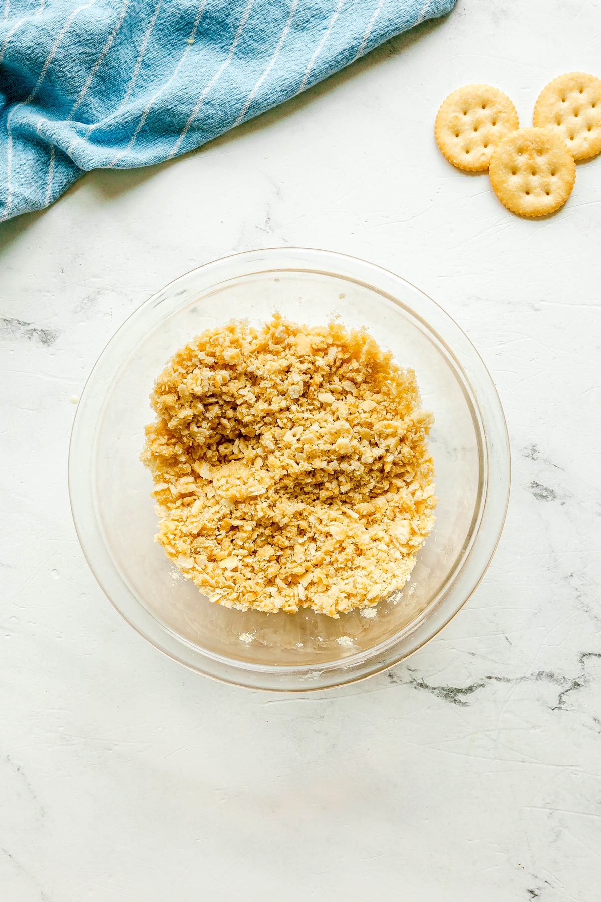 The first step in preparing Ritz Cracker Casserole is to use a clear bowl filled with crushed Ritz crackers and butter, sits on a white surface next to whole round crackers and a blue striped cloth.